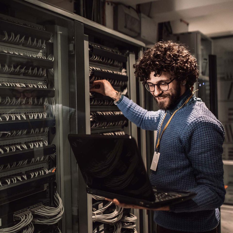 young IT professional wearing with curly brown hair and glasses checks cables in server room while looking at a laptop