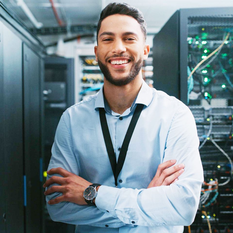 young IT professional smiles with arms folded at his waist in front of servers in server room