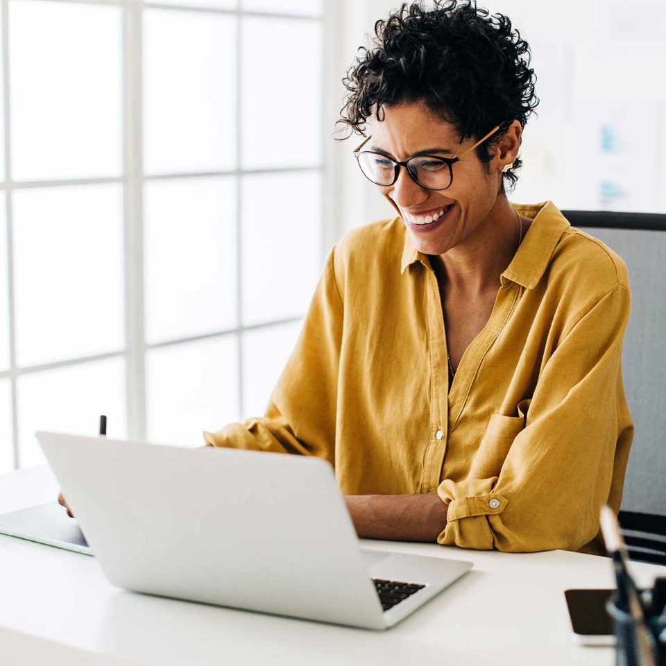 woman with glasses smiles as she works on a laptop in an office